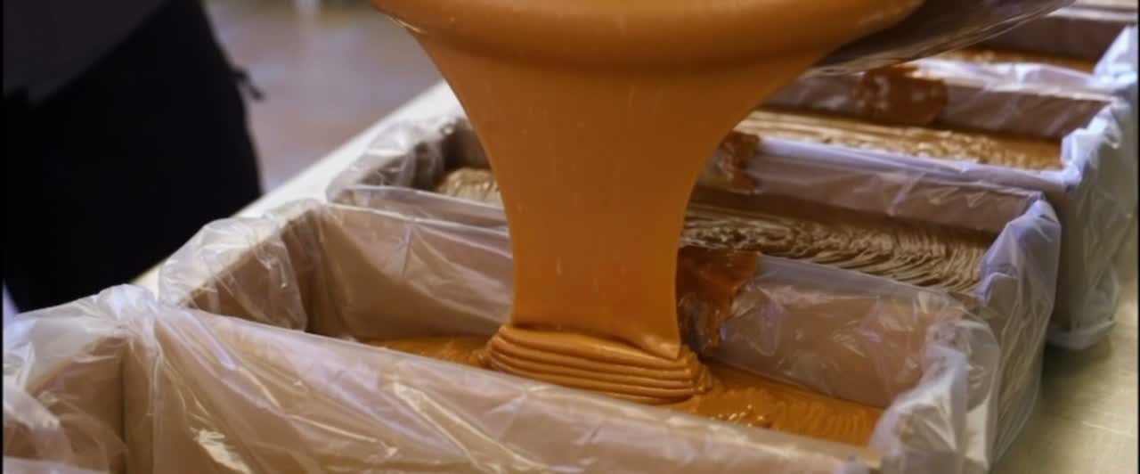 Molten fudge pouring in a thick ribbon into a lined loaf tin, with finished loaves behind it.